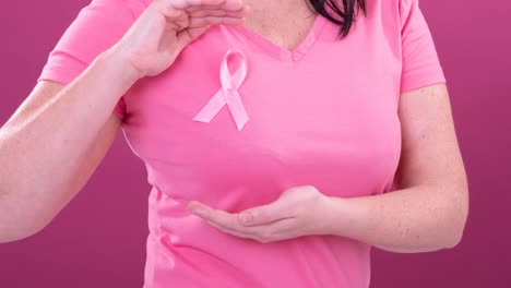 video of midsection of caucasian woman wearing pink cancer awareness ribbon, with pink background