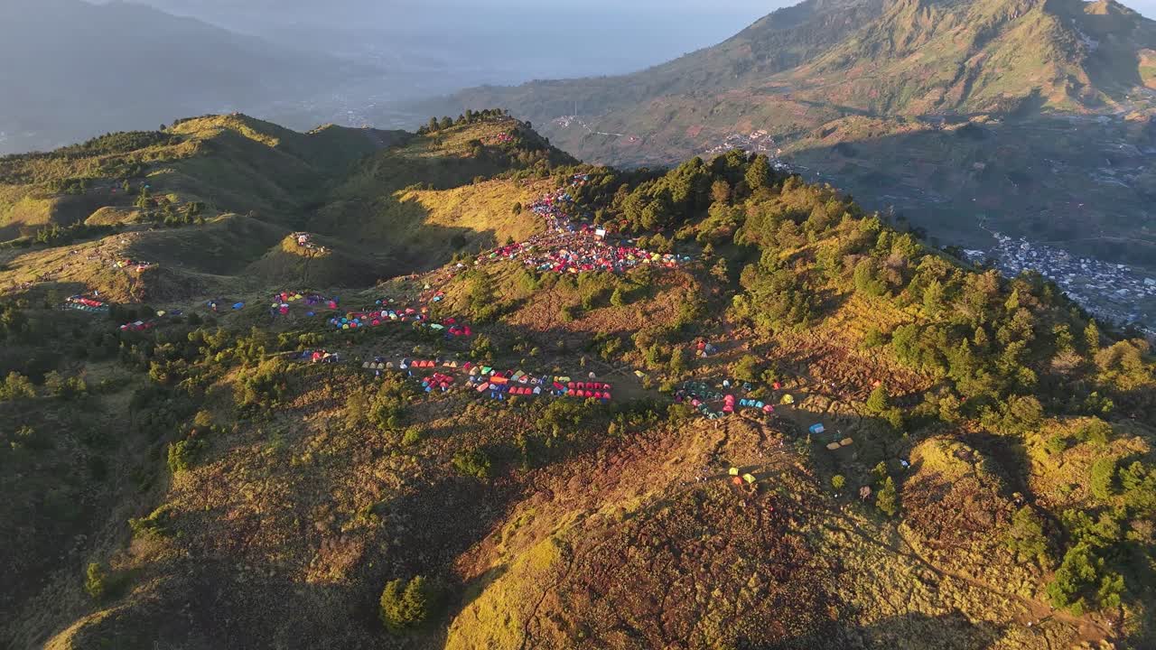 Scenic aerial panorama of hikers’ tents dotting a mountain summit, surrounded by green vegetation and valley views, evoking a sense of community and wilderness escape. Mount Prau, Indonesia