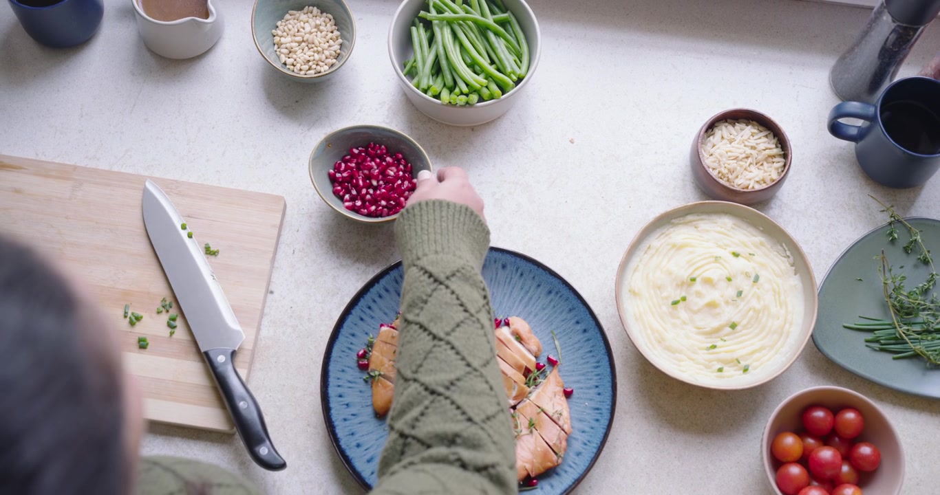 Two adults placing mug and chopping chives at counter, sprinkling pomegranate on blue plate chicken