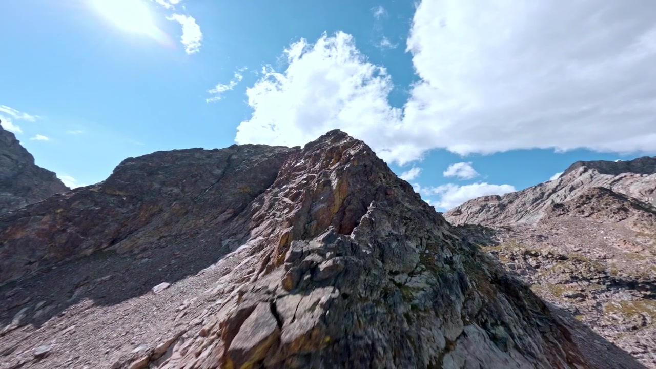 Drone FPV sfreccia lungo una cresta rocciosa esposta sotto il sole cocente di mezzogiorno, con pendii aridi e distanti e cielo luminoso tutt'intorno