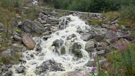 Braunes-Hochwasser-Strömt-Aus-Einem-Kleinen-Abflussrohr-Unterhalb-Der-Norwegischen-Straße-–-Statischer-Clip-Mit-Blick-Auf-Den-Fluss-Und-Den-Straßenzaun