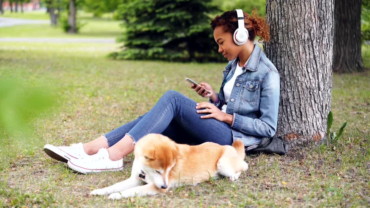 Attractive African American woman is listening to music with headphones and using smartphone sitting on grass in park while her dog is lying nearby eating grass.
