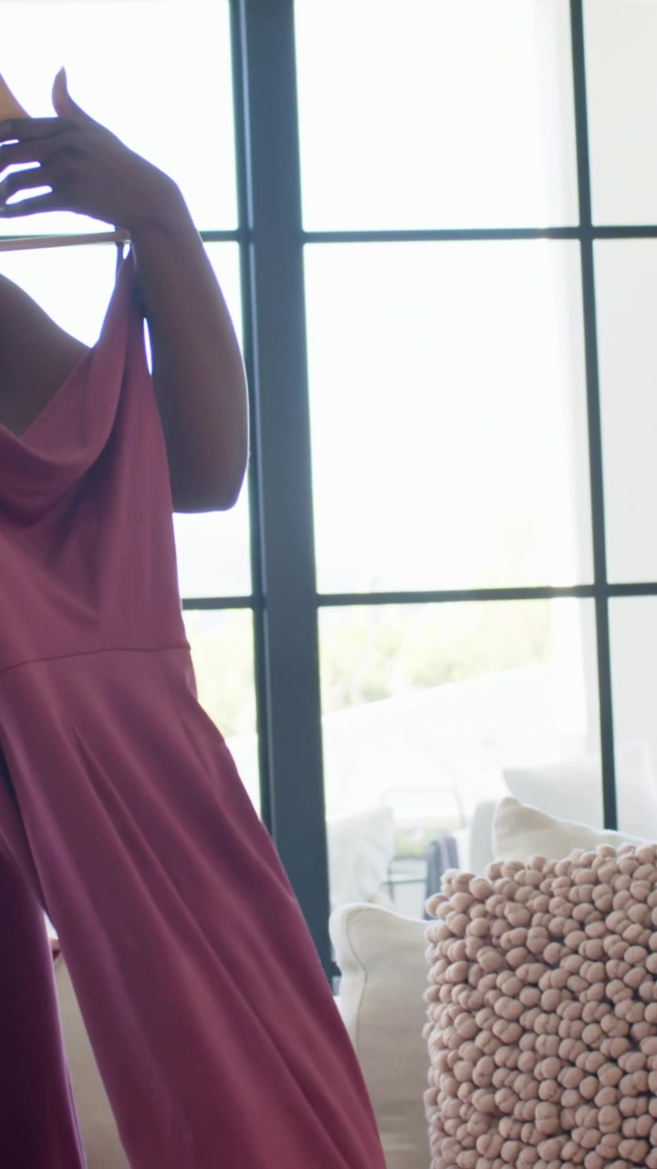 Video verticale: Donna in pantaloni che si alza, mostrando un vestito rosa sul divano per un parere, con un flûte di champagne