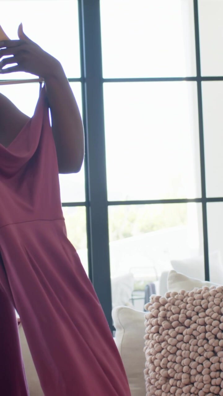 Vertical video: Rising woman in pants showing pink dress on couch for opinion with champagne flute