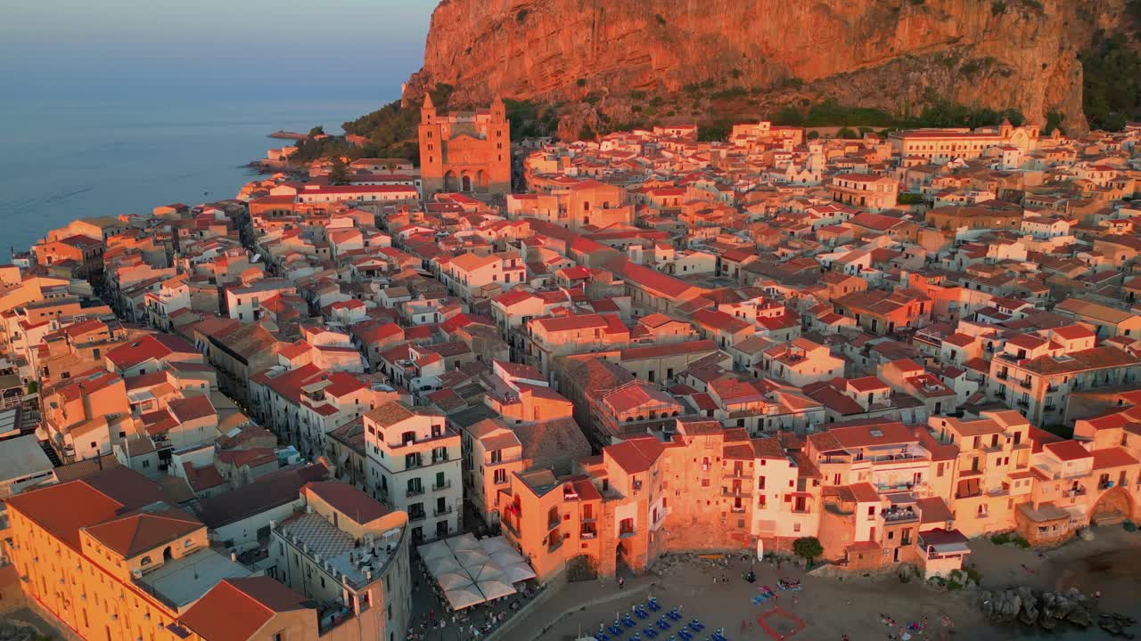 Luftfoto af Cefalù, der afslører en tæt, gammel by, sandstrand og roligt turkisblåt vand langs den sicilianske kyst under solnedgangen