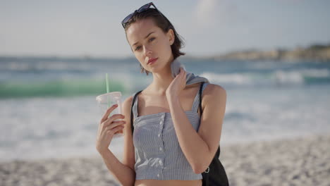 portrait of beautiful young woman on sunny beach looking at camera enjoying warm summer vacation day attractive caucasian female holding juice beverage fresh ocean seaside background