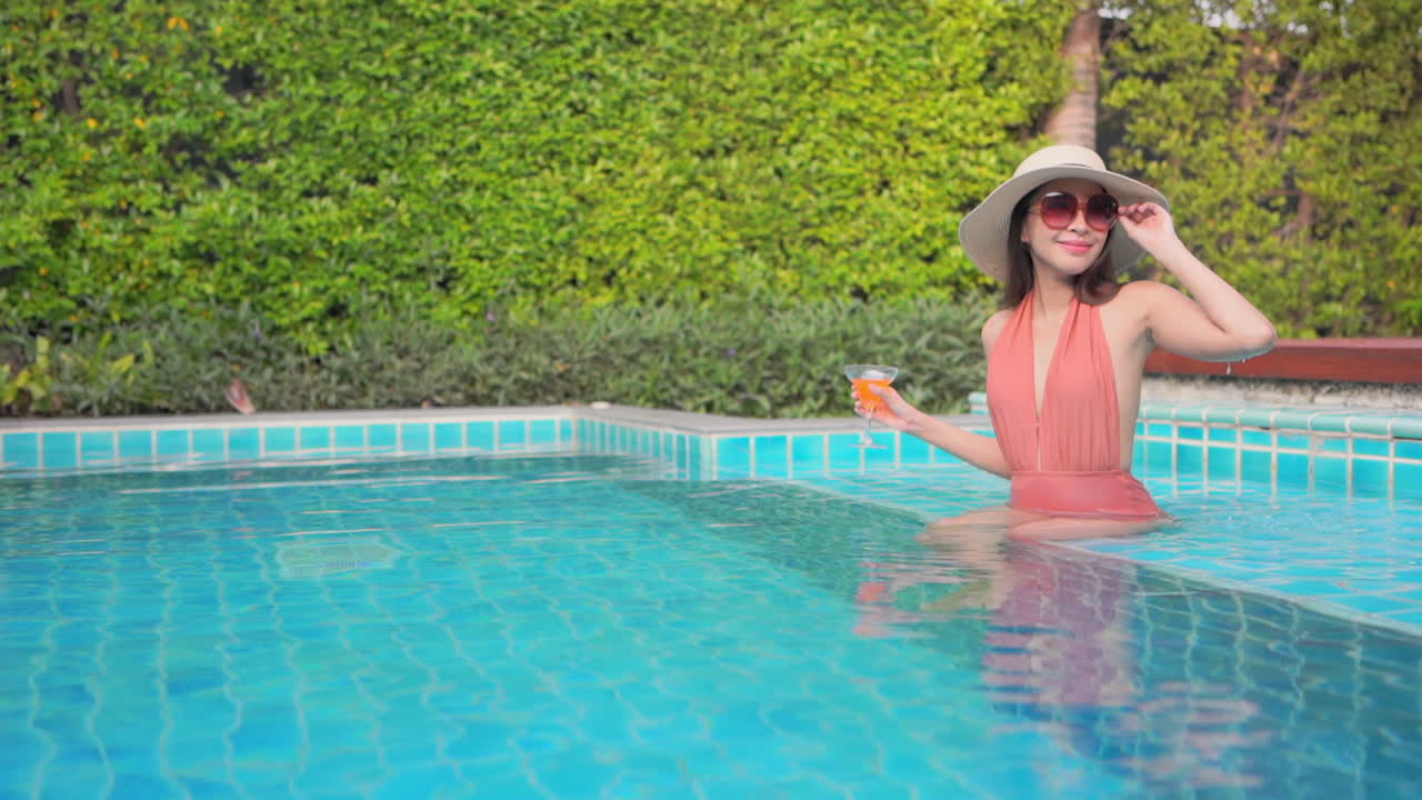 una mujer atractiva sentada en el escalón poco profundo de una piscina del complejo sostiene una bebida naranja colorida en una mano mientras ajusta sus gafas de sol con la otra