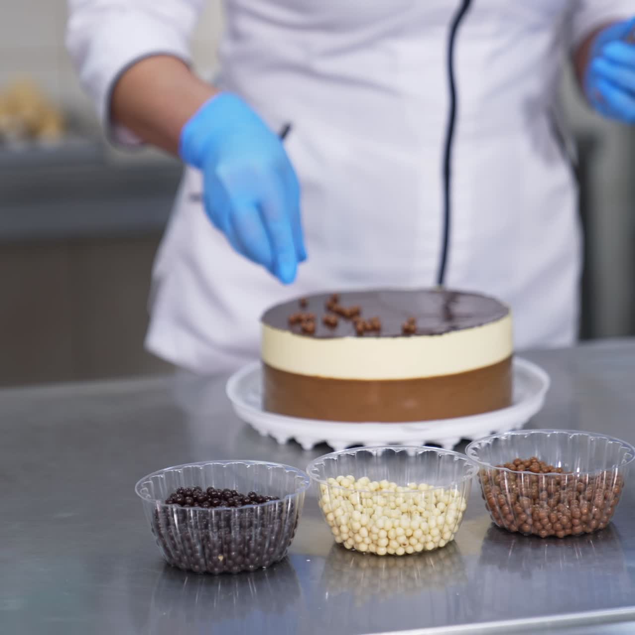 White and chocolate cake being decorated by the confectioner. Woman in gloves takes different candies from plastic cups and sprinkles them on top of the cake