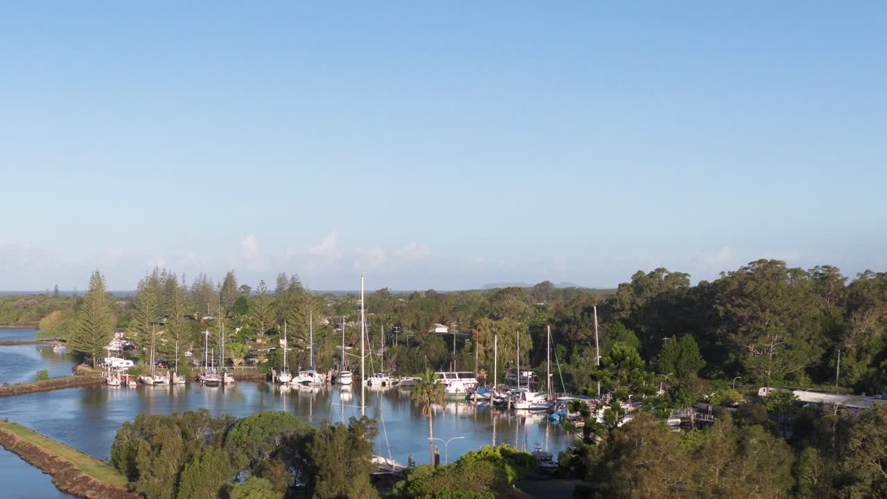 Aerial footage captures tranquil marina with boats and lush greenery under clear blue skies in Brunswick Heads, Australia