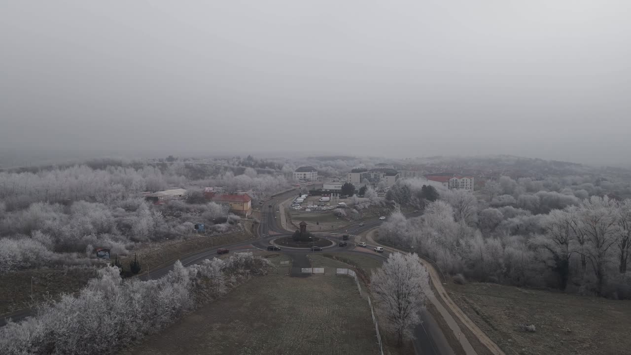 An aerial video of a roundabout surrounded by frost-covered trees and buildings in a winter landscape.