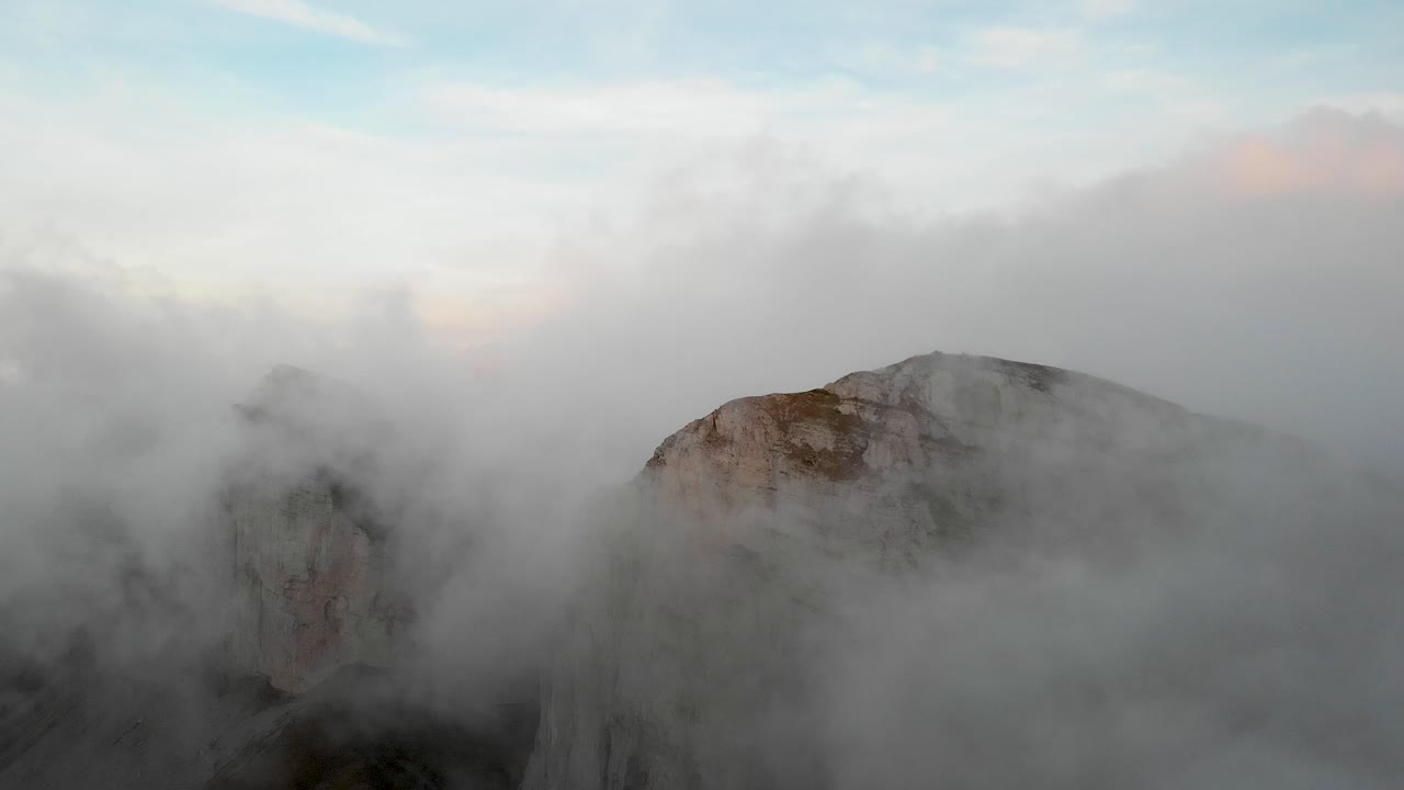 Aerial flyover through clouds with view of Tour d'A&iuml; and Tour de Mayen in Leysin, Vaud, Switzerland with a colorful alpenglow on the Swiss Alps in the background
