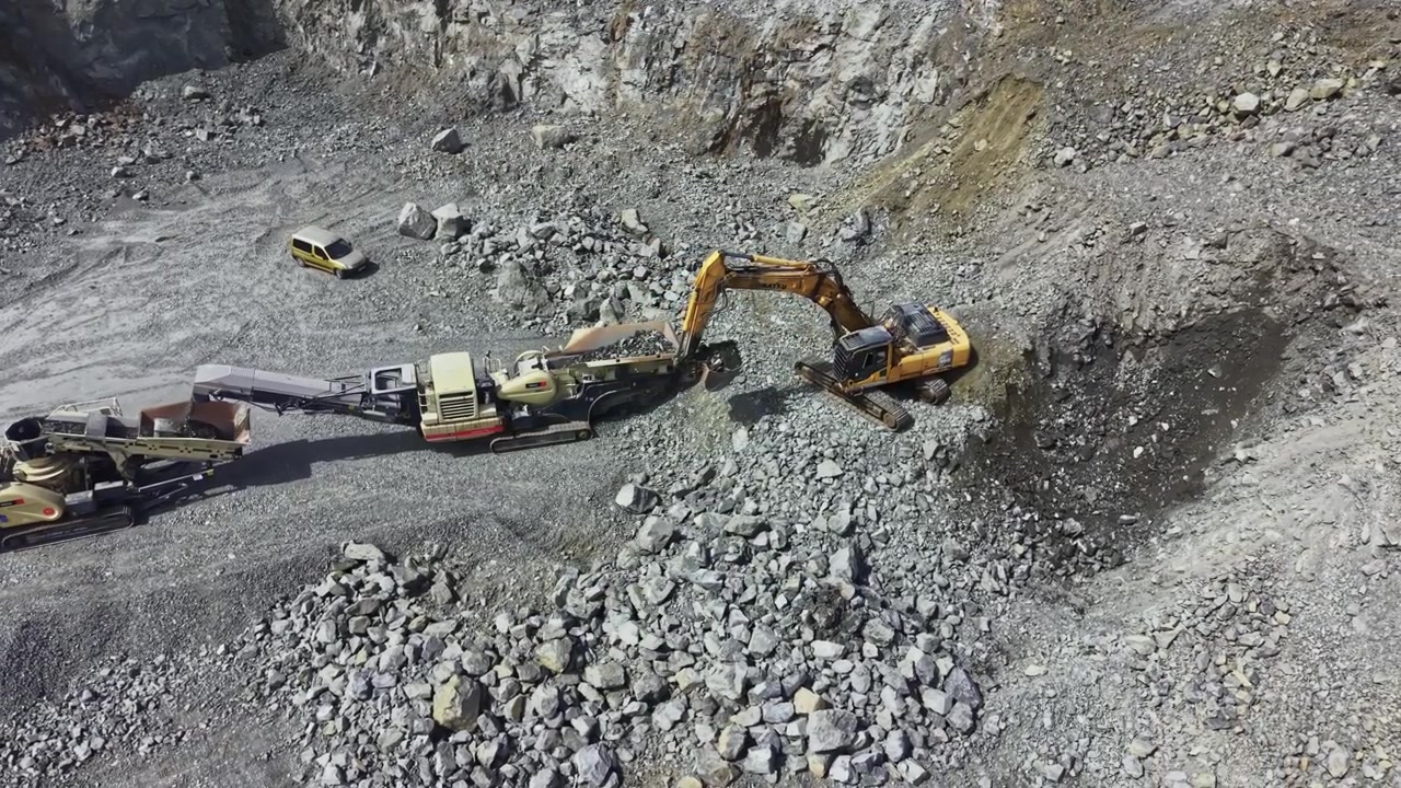Work occurs in a granite quarry with heavy machinery extracting stone and moving materials during a sunny day