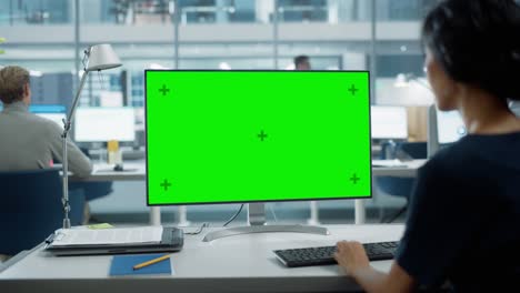 close up over the shoulder shot of a businesswoman working on desktop computer with chroma key green screen mock up display. digital projects manager typing data, using keyboard and mouse.