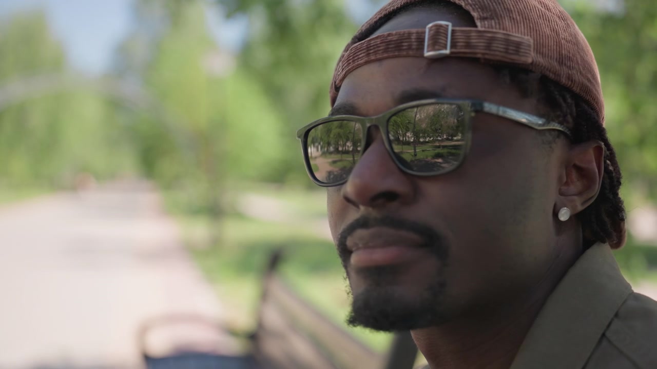 Close up of man wearing sunglasses and orange cap sitting on bench in bright daylight. scene features green trees blurred background with soft lighting capturing subject profile as man looks