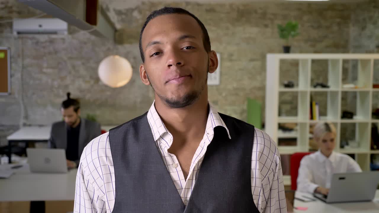 Young african businessman is standing and watching at camera in office, smiling, colleagues are working with laptops, work concept