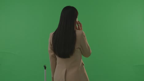 vista trasera mujer de negocios asiática viajera con equipaje hablando en teléfono inteligente mientras está de pie en el estudio de fondo de pantalla verde, durante la hora de la puesta del sol