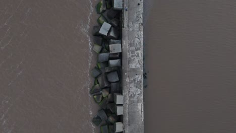 Top-down aerial view following the breakwater at Montevideo’s port, capturing the calm river-sea figure either a fisherman or a visitor walking toward the lighthouse at the end of the structure