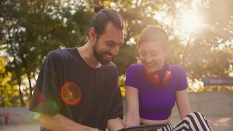 Close-up-shot-of-a-brunette-guy-in-a-gray-T-shirt-and-a-girl-with-a-short-haircut-in-a-purple-top-looking-at-something-on-the-tablet-screen-in-the-summer-park