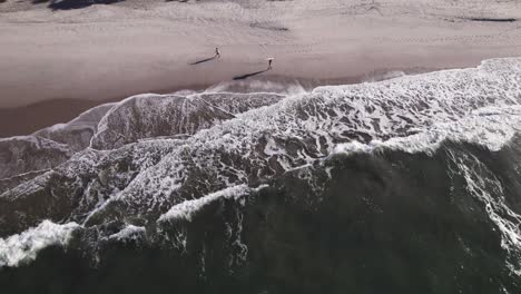 Un-Dron-Estático-Muestra-La-Orilla-Cuando-Dos-Hombres-Se-Cruzan-Cerca-De-Las-Olas,-Uno-Con-Su-Tabla-De-Surf-Sobre-La-Cabeza