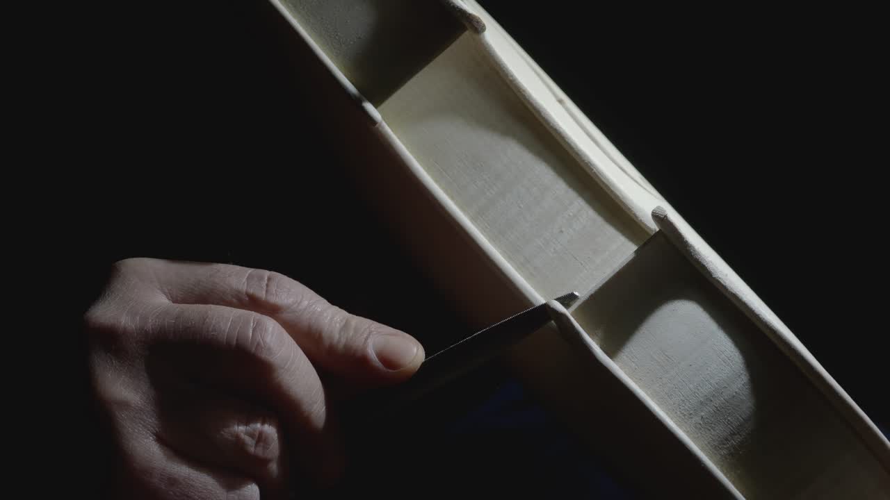 Extreme close up macro shot in slow motion of hand of master lute maker smoothing and shaping corner edges near the center bout with raw rasp to new unpainted classical violin