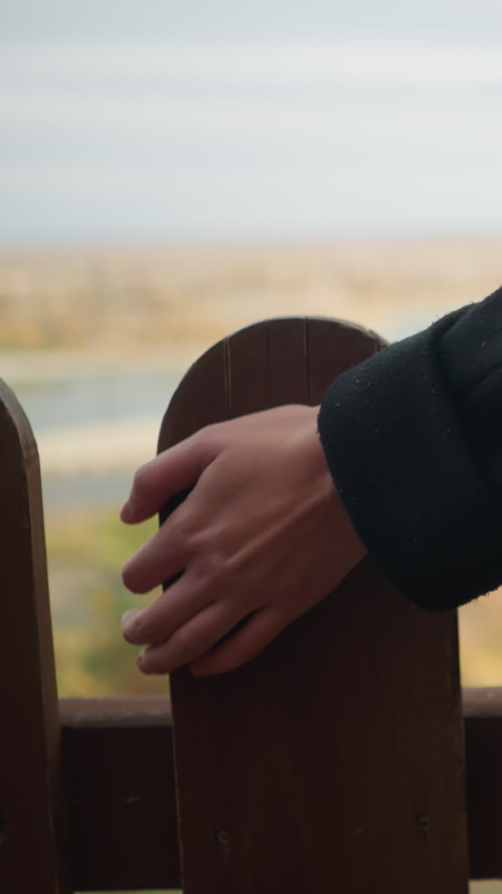 primer plano de la mano de una dama en un abrigo negro tocando delicadamente una valla de madera, con árboles de enfoque suave en la distancia creando un telón de fondo sereno