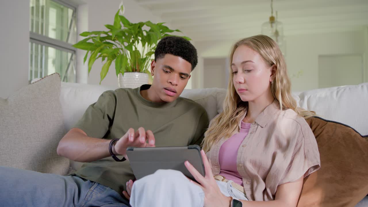Diverse couple sitting on sofa in living room, woman explaining app on tablet, man testing screen