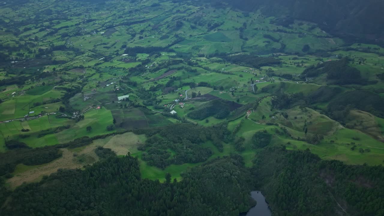 Drone advancing over farmland near Zipaquirá, showing patchwork fields, scattered rural houses and a small section of a forested lake at the edge of the frame