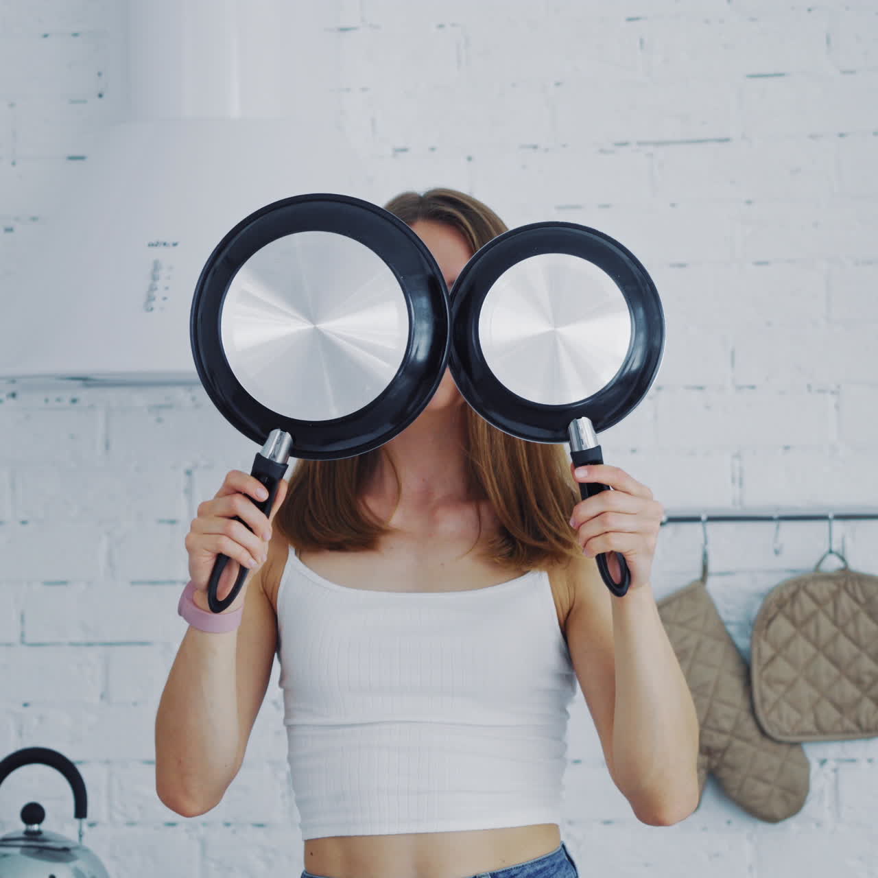 Young female with frypan in the kitchen. Attractive housewife holding two new nonstick pans and closing her face by them. Happy woman in the modern kitchen.