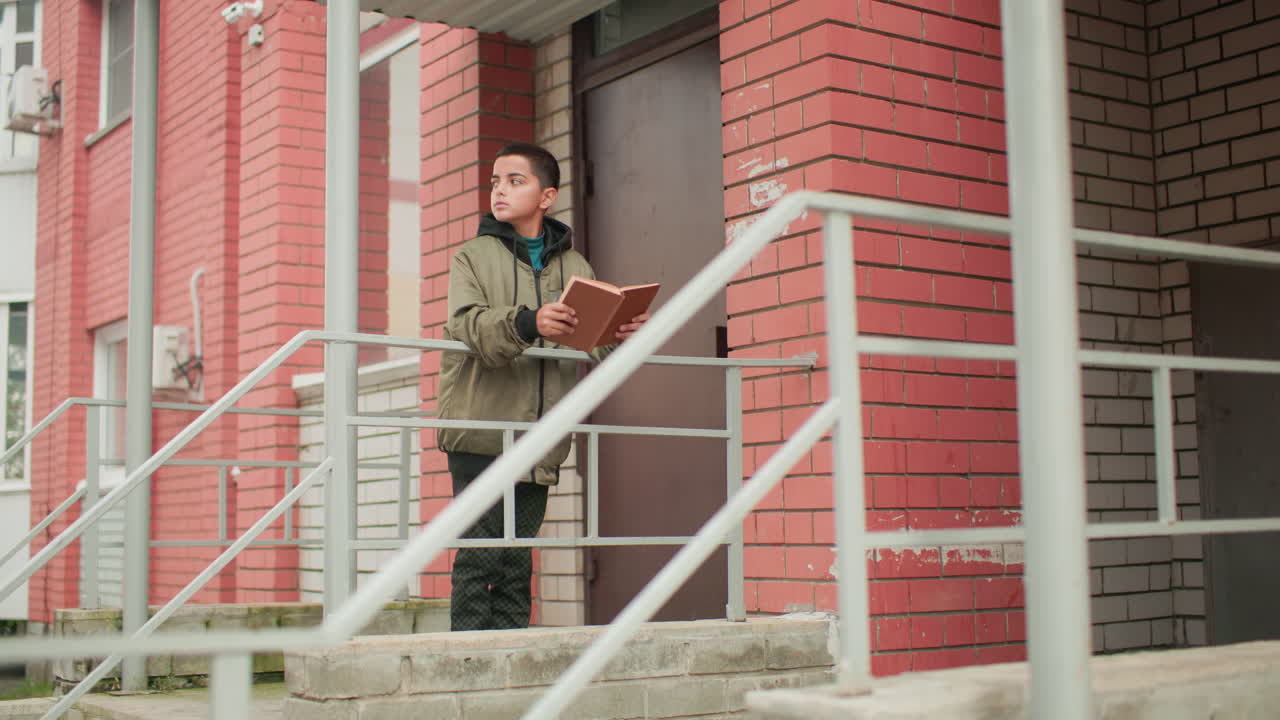 Boy in hooded jacket stands near red brick building holding open brown book, studying with focus as he briefly glances back before continuing to read attentively outdoors