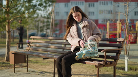 a girl wearing a peach jacket, grey inner shirt, and black trousers is seen walking and sitting on a park bench, placing her rollerblades beside her. a moment of relaxation