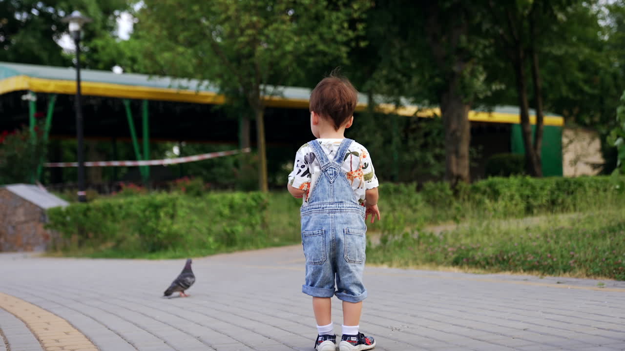 Cute baby boy in a t-shirt and jeans romper is on the walk. Little kid walks by the paved road outdoors.