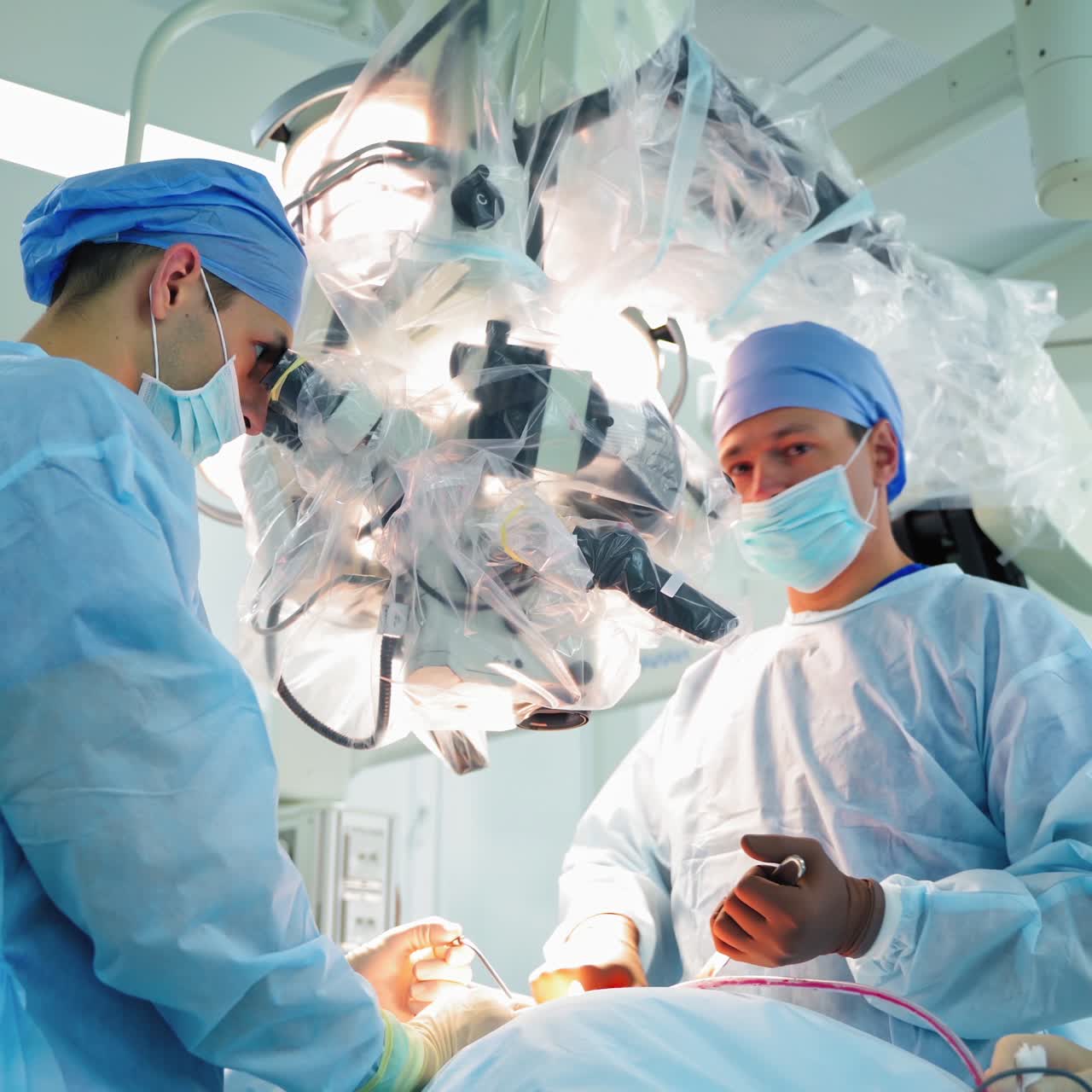 Teamwork of neurosurgeons. Medical specialists look into the microscope while doing a surgery in the operating room. Assistant male helps doctors in hospital.