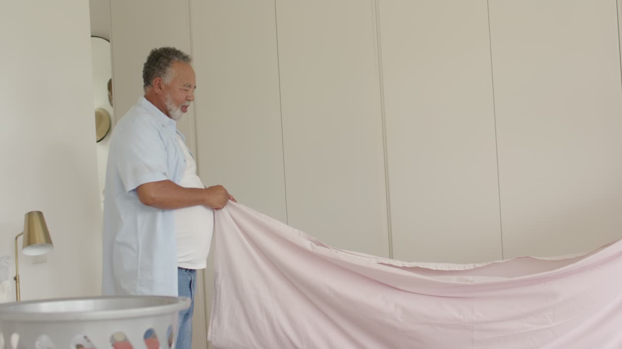 Pareja madura sacudiendo y extendiendo una sábana rosa sobre la cama después de la colada, con una cesta, alisando el colchón.