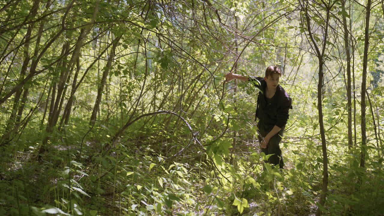 Tourist moves backward through dense green forest while adjusting shoulder bag and holding branch to clear path, surrounded by overgrown foliage and sunlight filtering through trees