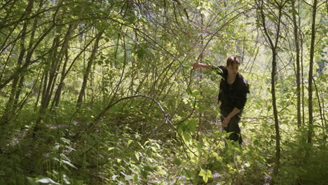 tourist moves backward through dense green forest while adjusting shoulder bag and holding branch to clear path, surrounded by overgrown foliage and sunlight filtering through trees