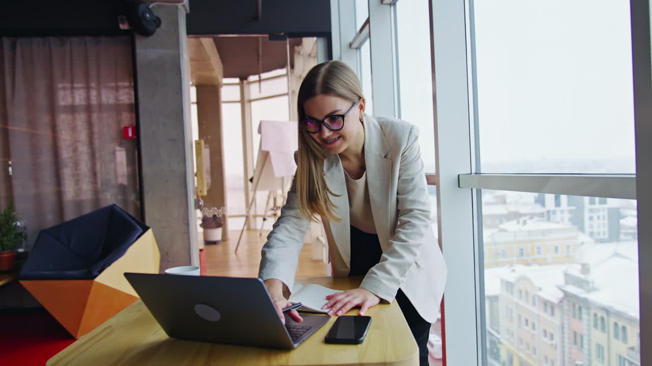 Businesswoman standing over the table and looking at laptop. Happy woman typing on computer and smiling. Big light office backdrop.