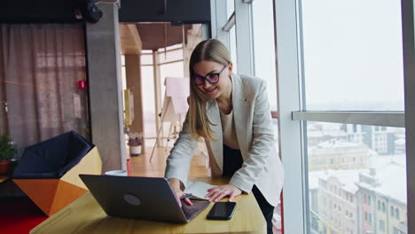 businesswoman standing over the table and looking at laptop. happy woman typing on computer and smiling. big light office backdrop.