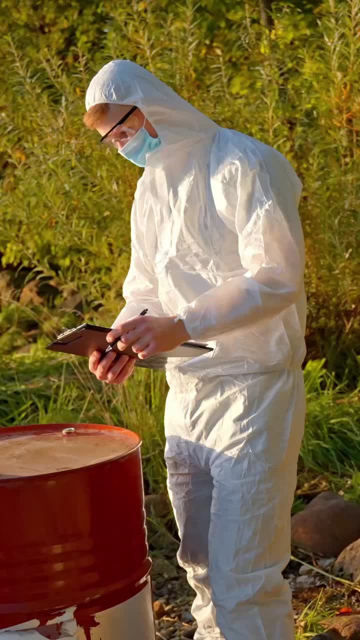 Scientist in white hazmat suit takes notes beside oil drum at polluted river shoreline, vertical