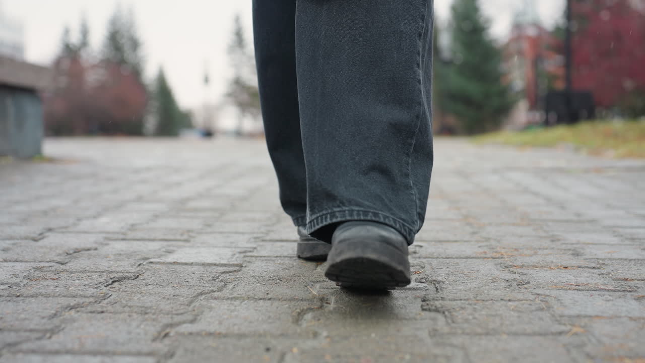 Close up of person's black trousers and black boots walking slowly toward camera on paved urban path, face not visible, capturing detail of steps and movement on cloudy chilly autumn day outdoors