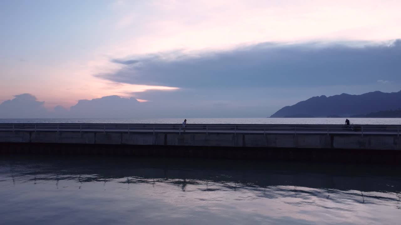 Incredibile vista aerea di una figura in controluce che cammina lungo un molo di cemento sull'oceano durante un tramonto tranquillo e nuvoloso, con l'acqua calma che riflette il cielo crepuscolare colorato.