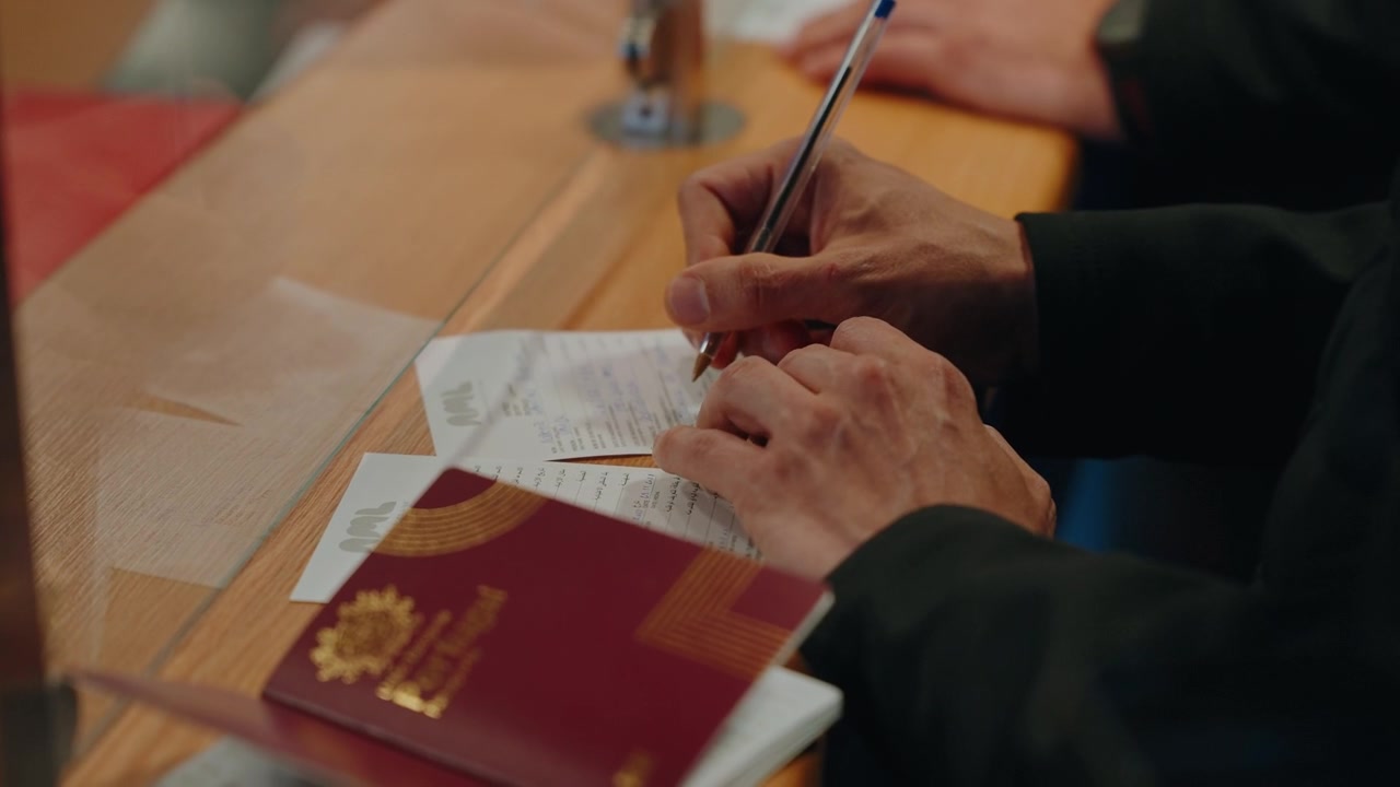 Ruce podepisující příjezdový imigrační formulář vedle pasu a cestovních dokladů u hraničního pultu