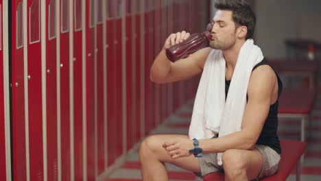 Closeup-exhausted-man-sitting-in-dressing-room-with-sport-bottle.