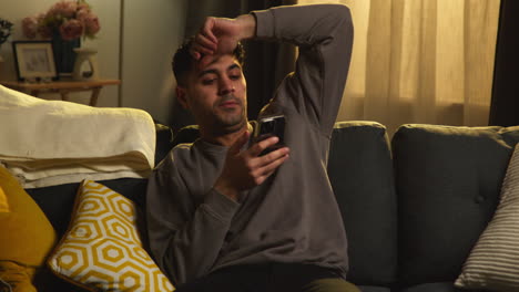 young man spending evening at home sitting on sofa with mobile phone scrolling through internet or social media