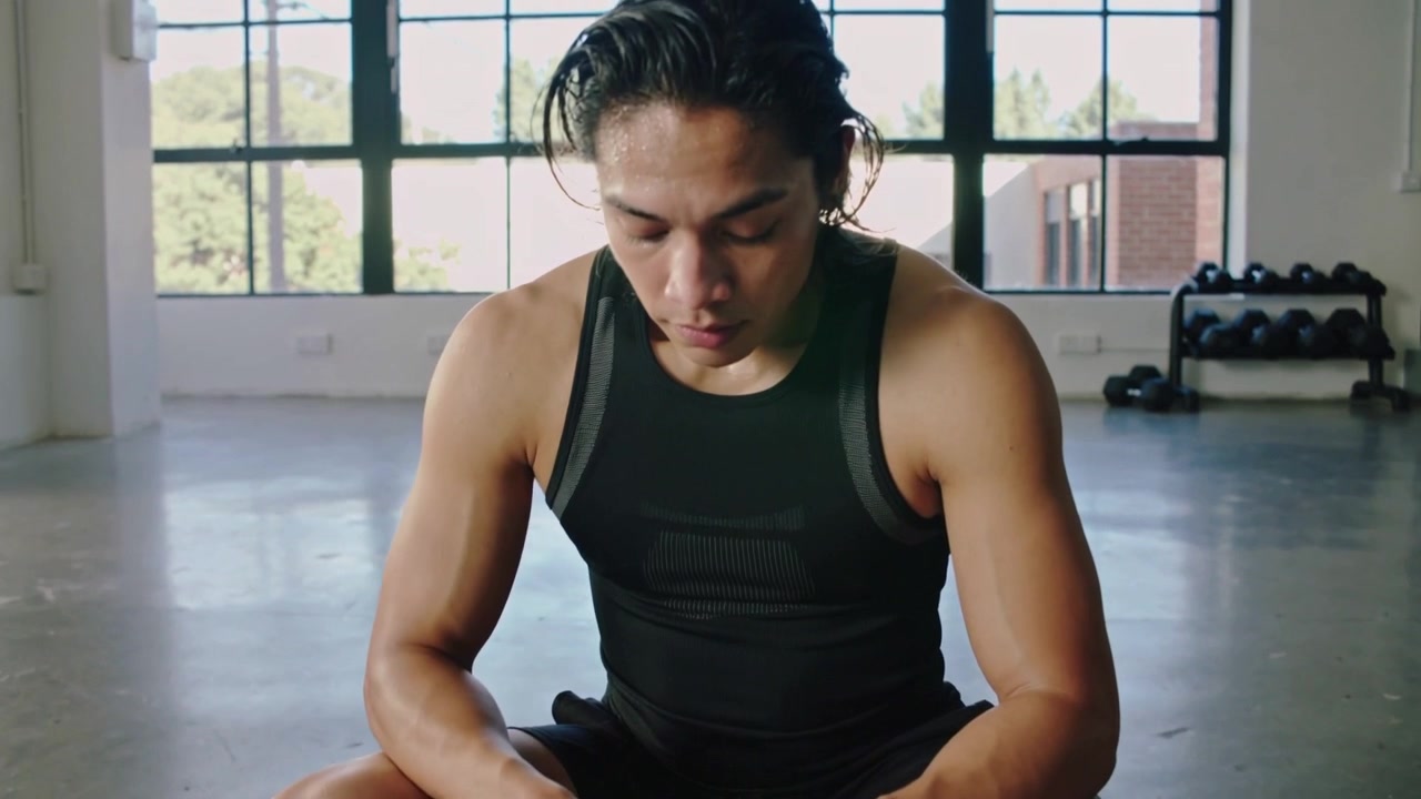 Menyelesaikan set, pria mengenakan atasan tanpa lengan dan celana pendek menyeka keringat, duduk di gym dengan rak dumbel.