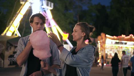 una joven pareja hipster está pasando el tiempo juntos en el parque de atracciones por la noche. alimentándose mutuamente con algodón de azúcar rosa. vista frontal