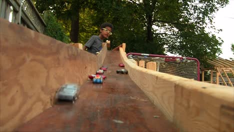 a young boy is racing his diecast hot wheels on a homemade wooden racetrack in his backyard