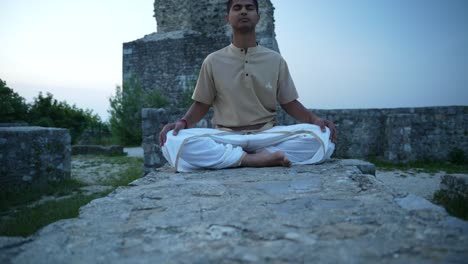 young indian man opening his chakras with deep meditation and hatha yoga while sitting on stone wall of castle ruins on top of the hill at sunrise