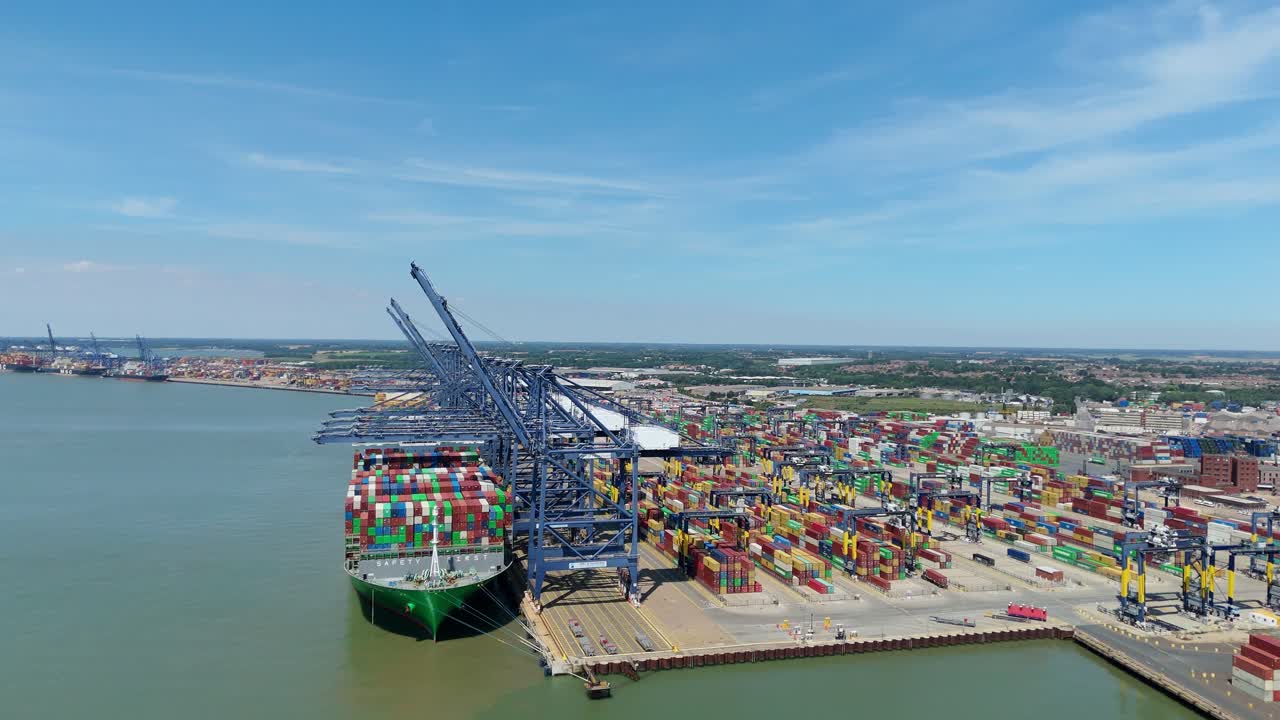 Aerial drone footage of a large container ship being loaded with freight by heavy cranes at Felixstowe port harbour, UK sunny summer day busy import and export