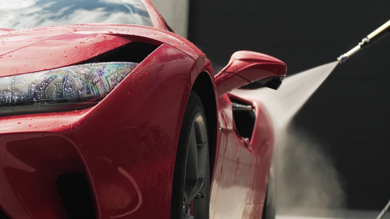 Red sports car being washed with high-pressure water, creating mist and shine on a sunny day