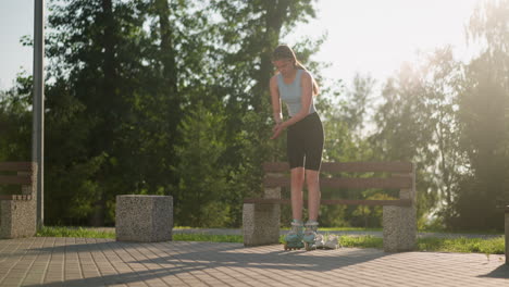 young woman attempting to balance as she stands up from bench while wearing roller skates in park, with white sneakers under bench and sunlight illuminating green trees in background